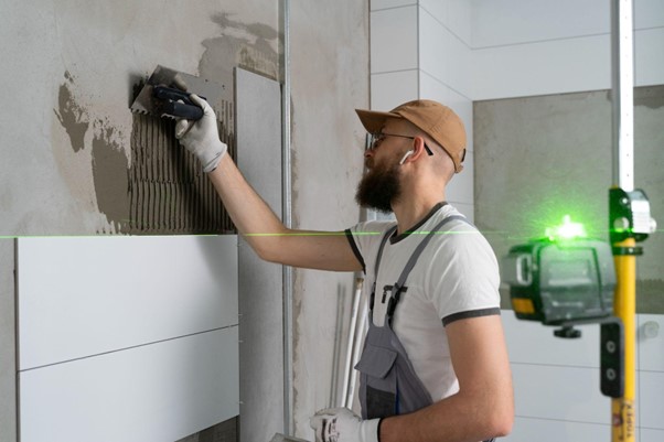 Řemeslník pokládá dlaždice na zeď a používá při tom stěrku. Laserové zařízení mu pomáhá udržet rovnou linii.