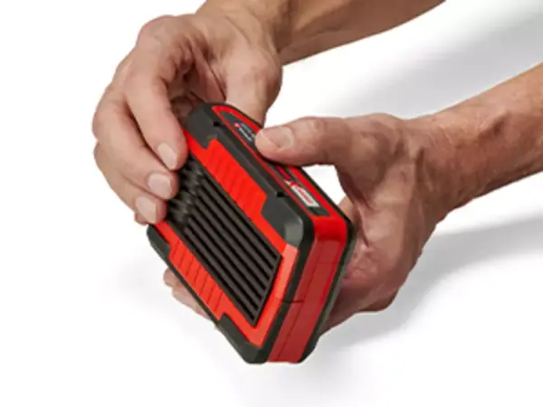Dvě ruce drží akumulátor Einhell Power X-Change za vroubkovanou úchopovou drážku; viditelné je robustní pogumované pouzdro.